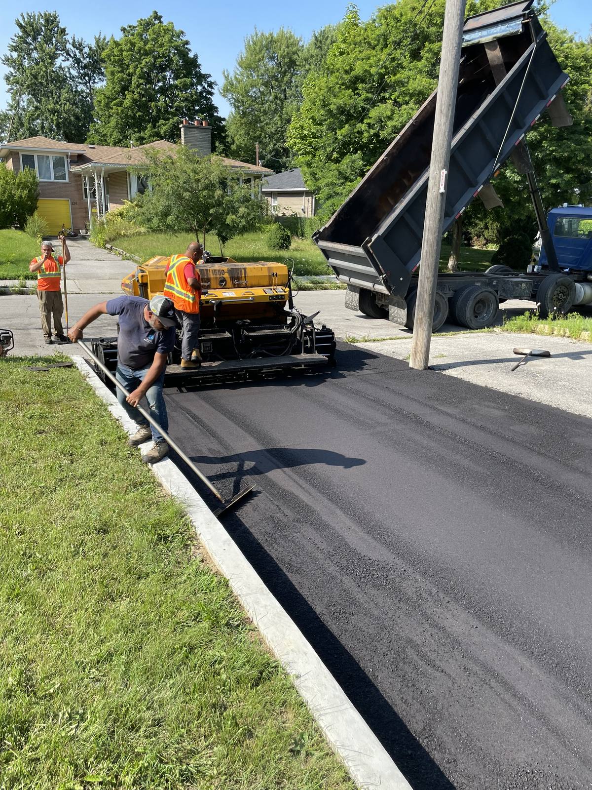 Crew machine-paving a residential driveway, screeding fresh asphalt against a new concrete curb — Berm Inc.